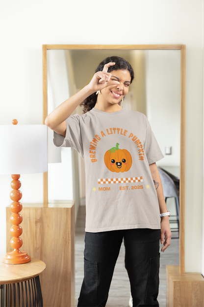 Brewing A Little Pumpkin T-Shirt in sand color worn by a cheerful young woman, featuring an adorable pumpkin with a pacifier and the text "Brewing a Little Pumpkin, Mom Established 2025." Cute and cozy shirt perfect for announcing a pregnancy during Halloween and makes a charming gift for expecting parents. Ideal for festive gatherings and family photos