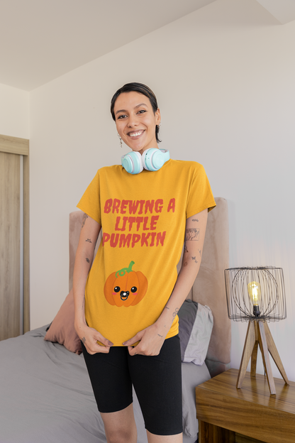 Gold color t-shirt featuring red text "Brewing A Little Pumpkin," perfect for announcing a pregnancy during Halloween, worn by a smiling woman.