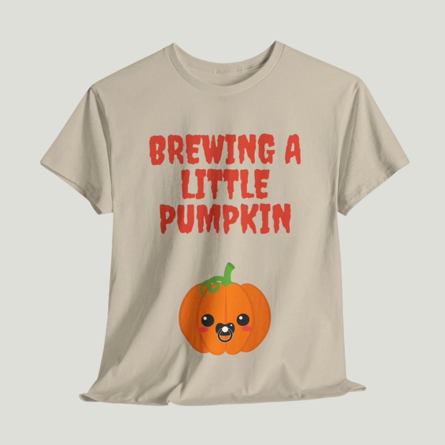 sand t-shirt featuring red text "Brewing A Little Pumpkin," perfect for announcing a pregnancy during Halloween, worn by a smiling woman.