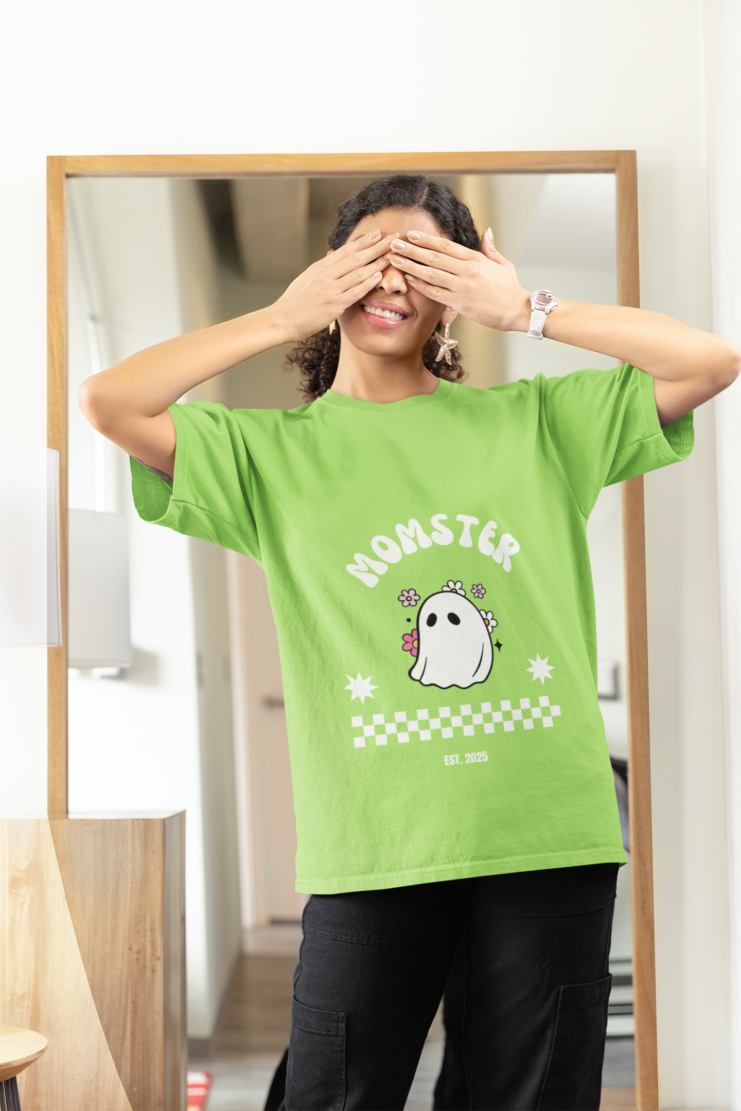 Woman wearing a Momster T-Shirt in green color with a cute ghost graphic and the text 'Established 2025,' showcasing a playful and festive way to announce a pregnancy during Halloween.